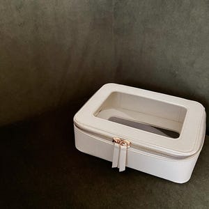 May include: A white rectangular cosmetic bag with a clear top panel. The bag has a silver zipper and a gold-coloured zipper pull. The bag is on a dark green surface.