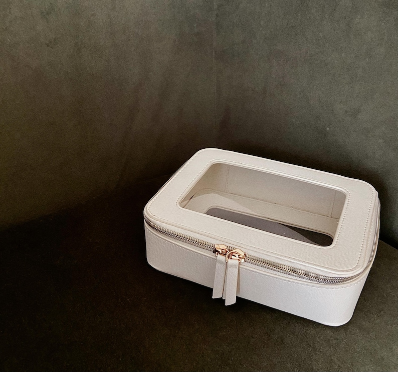 May include: A white rectangular cosmetic bag with a clear top panel. The bag has a silver zipper and a gold-coloured zipper pull. The bag is on a dark green surface.