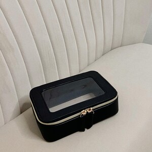 May include: A black rectangular cosmetic bag with a clear top panel. The bag has a gold zip and a small pull tab. The bag is on a white, ribbed chair.