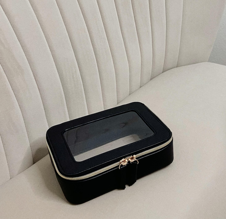 May include: A black rectangular cosmetic bag with a clear top panel. The bag has a gold zip and a small pull tab. The bag is on a white, ribbed chair.