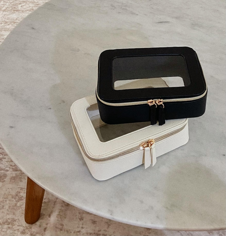 May include: Two square cosmetic cases, one black and one white, with clear tops and gold zips. The cases are stacked on a marble-topped table. The cases are designed for storing makeup and beauty products.