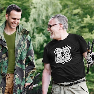 May include: Two people are smiling and laughing outdoors. One wears a camouflage jacket and olive green shirt, the other a black t-shirt with a "Forest Service Sasquatch" logo. They are holding fishing gear and a net.