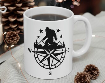 May include: White ceramic mug with a black silhouette of Bigfoot standing on a compass rose with stars. The text "W" and "E" are on the compass rose.