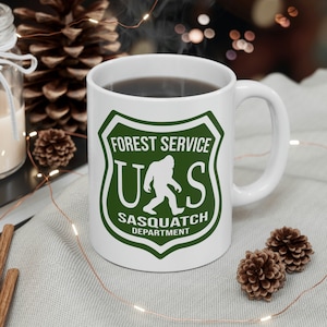May include: White ceramic mug with a green forest service logo. The logo features a silhouette of Bigfoot walking and the text "US Forest Service Sasquatch Department".