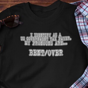 May include: Black t-shirt with a white graphic that says "I identify as a US Government Tax Payer. My pronouns are... Bent/Over."