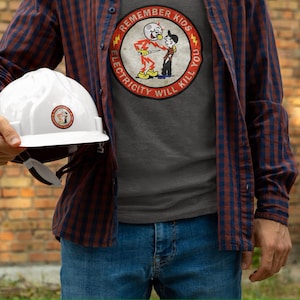 May include: A man wearing a gray t-shirt with a red circle graphic that says "Remember Kids Electricity Will Kill You" and a white hard hat. The man is wearing a red and blue plaid shirt over the t-shirt.