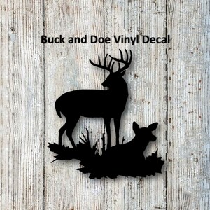 May include: Black vinyl decal featuring a buck and doe silhouette standing on a bed of leaves and grass. The text "Buck and Doe Vinyl Decal" is above the image. The background is a weathered, light-colored wood.