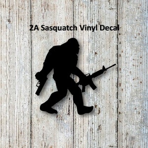 May include: Black vinyl decal depicting a Bigfoot silhouette armed with two guns. The text "2A Sasquatch Vinyl Decal" is above the image. The background is a light-coloured, weathered wooden surface.