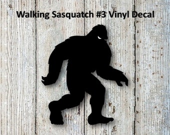 May include: Black vinyl decal of a walking Sasquatch silhouette. The text "Walking Sasquatch #3 Vinyl Decal" is printed above the image. The background is a light-colored wood grain.