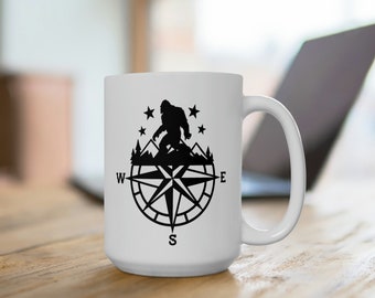 May include: White ceramic mug with a black silhouette of Bigfoot walking on a mountain range with stars above. The mountain range is on top of a compass with the letters N, S, E, and W.