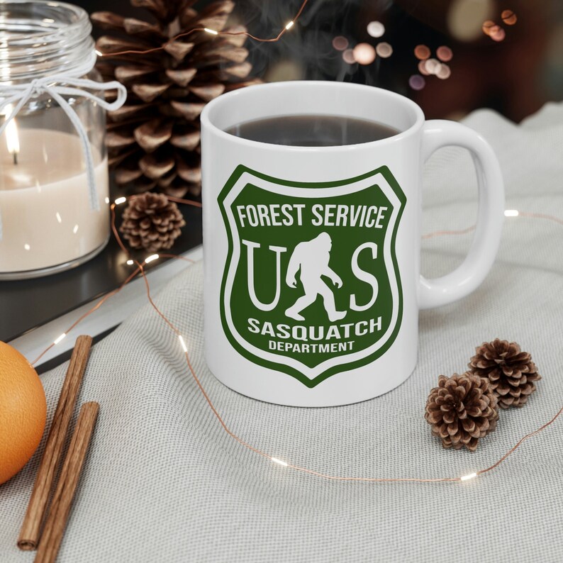 May include: White ceramic mug with a green forest service logo. The logo features a silhouette of Bigfoot walking and the text "US Forest Service Sasquatch Department".