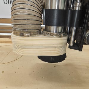 May include: Close-up of a wood dust collection system for a CNC router. The system includes a flexible hose, a metal clamp, and a brush.