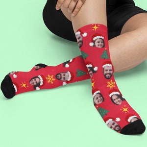 May include: Red Christmas socks with a repeating pattern of faces wearing Santa hats, green Christmas trees, gold stars, and snowflakes. The socks have black heels and toes.