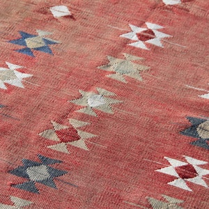 Op de afbeelding: Een rood tapijt met een geometrisch patroon in tinten wit, blauw en beige. Het patroon bestaat uit herhalende ruitvormen.