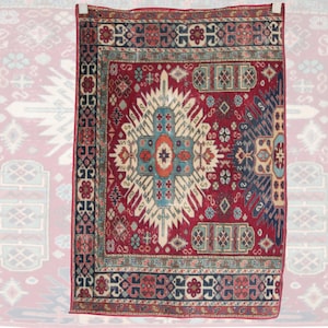 Puede incluir: Una alfombra rectangular con un fondo rojo intenso y un medallón geométrico central en azul, naranja y crema. La alfombra está enmarcada por un borde ancho con motivos florales y geométricos repetidos en azul marino, rojo y crema.