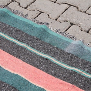 May include: A striped rug with teal, black, and pink stripes. The rug is lying on a gray concrete surface.