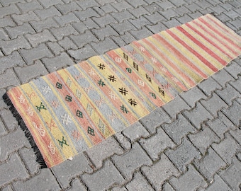 Può includere: Un tappeto runner rettangolare tessuto con un design a righe colorate. Il tappeto presenta strisce alternate rosse, gialle e grigie, con motivi geometrici verdi e marroni. Il tappeto è posizionato su una superficie di mattoni grigi.