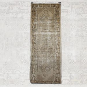 May include: A rectangular runner rug featuring a faded beige and tan color palette. The rug displays a central medallion design surrounded by an intricate geometric border pattern with repeating motifs. The edges are finished with short fringe.