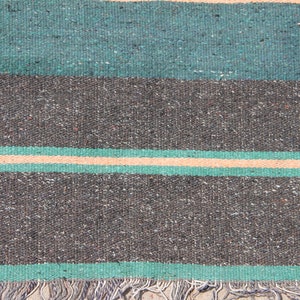 May include: A close-up of a woven rug with a striped pattern in shades of green, gray, and beige. The rug has a fringe border.