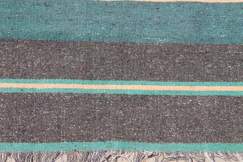 May include: A close-up of a woven rug with a striped pattern in shades of green, gray, and beige. The rug has a fringe border.
