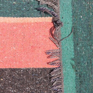 May include: Close-up of a woven rug with a green, pink, and black striped pattern. The rug has a fringe on the right side.