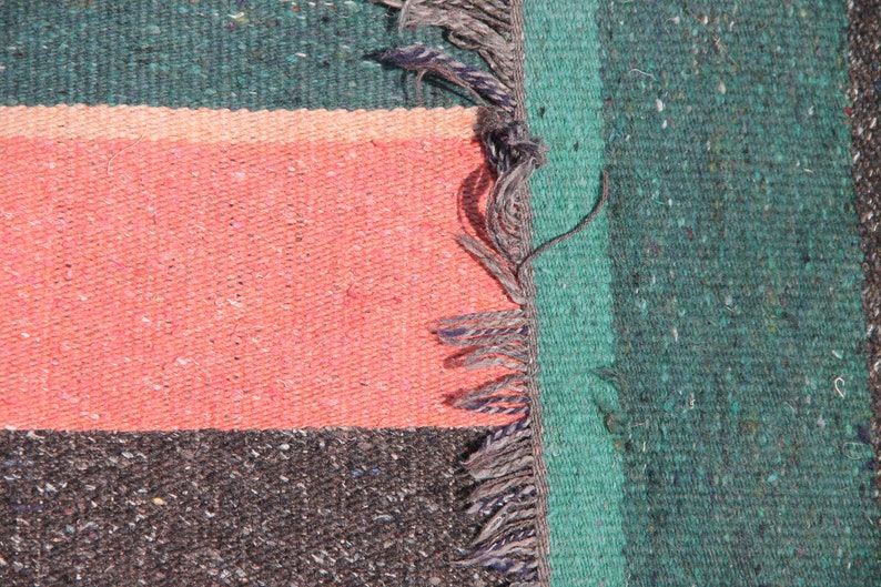 May include: Close-up of a woven rug with a green, pink, and black striped pattern. The rug has a fringe on the right side.