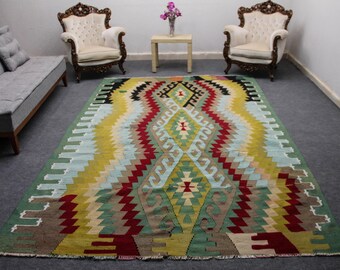 May include: A colorful, hand-woven rug with a geometric pattern in shades of red, green, yellow, and blue. The rug is laid out on a gray floor in a room with two white chairs and a gray sofa.
