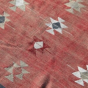 Op de afbeelding: Een rood geweven tapijt met een geometrisch patroon van witte en grijze sterren. Het tapijt heeft een verweerde, vintage uitstraling.
