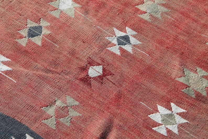 Op de afbeelding: Een rood geweven tapijt met een geometrisch patroon van witte en grijze sterren. Het tapijt heeft een verweerde, vintage uitstraling.