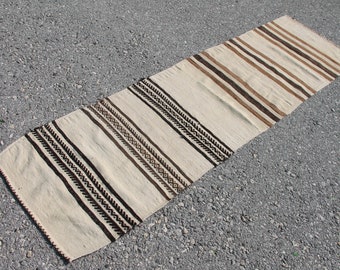 May include: A long, narrow, off-white rug with brown and black stripes and geometric patterns. The rug is woven with a simple, rustic design.