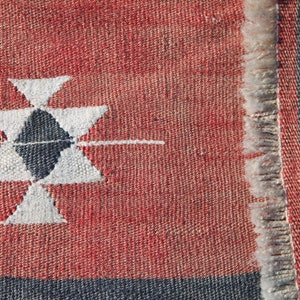 Op de afbeelding: Close-up van een rood en blauw geweven tapijt met een wit geometrisch patroon. Het tapijt heeft een franje rand.