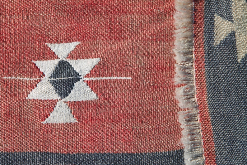 Op de afbeelding: Close-up van een rood en blauw geweven tapijt met een wit geometrisch patroon. Het tapijt heeft een franje rand.