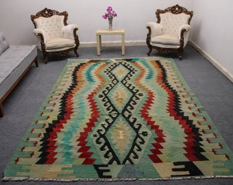 May include: A colorful vintage rug with a geometric pattern in shades of red, blue, green, and black. The rug is laid out on a gray floor in a room with two white chairs and a small table.