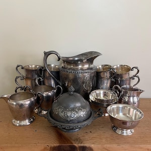 May include: An assortment of antique silver serving pieces, including a large pitcher, creamers, sugar bowls, and small bowls. The pieces have ornate detailing and are arranged on a wooden surface. The silver has a tarnished appearance, giving it a vintage look.