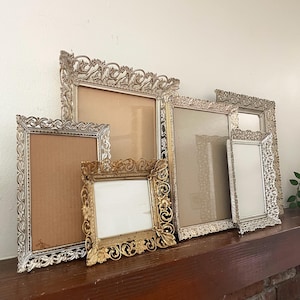 May include: A collection of ornate picture frames in various sizes. The frames are made of metal and feature intricate, scrolling designs. Some frames have a silver finish, while others are gold. The frames are arranged on a wooden surface.