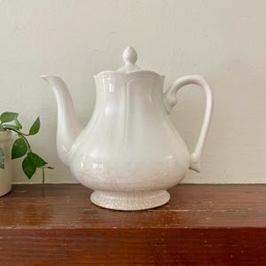 May include: A white ceramic teapot with a lid and a curved handle. The teapot has a bulbous body and a spout. The base of the teapot is slightly cracked. The teapot is sitting on a wooden surface.