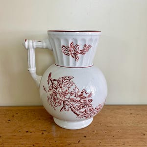 May include: White ceramic pitcher with a red floral design. The pitcher has a round body and a flared top. The handle is attached to the side of the pitcher and has a decorative knob.