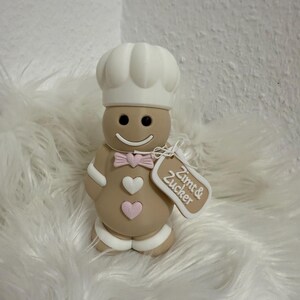 Könnte beinhalten: Lebkuchenmann-Figur mit Kochmütze und Fliege. Die hellbraune Figur hat weiße Zuckergussdetails, ein lächelndes Gesicht und zwei rosa Herzen. Ein Anhänger trägt die Aufschrift "Zimt & Zucker".