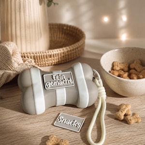 Könnte beinhalten: Ein grau-weißer Hundeknochen-förmiger Leckerli-Spender mit den Texten "Fein gemacht" und "Snacks". Ein weißes Seil ist befestigt. Hundeleckerlis befinden sich in einer Schüssel und sind auf einer Holzoberfläche verstreut.