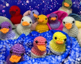 Op de afbeelding: Een verzameling kleurrijke gehaakte vogelversieringen in verschillende kleuren, waaronder paars, roze, geel, wit en groen. De versieringen zijn gerangschikt op een blauwe stoffen achtergrond met een glinsterende blauwe achtergrond.
