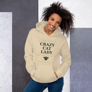 Może przedstawiać: Beżowa bluza z kapturem z przednią kieszenią i kapturem ze sznurkiem. Tekst "CERTIFIED CRAZY CAT LADY AND PROUD OF IT" jest nadrukowany z przodu w kolorze zielonym i brązowym. Poniżej znajduje się zielony odcisk łapy kota.