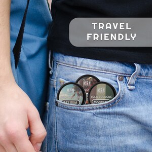 May include: Three small round containers of solid cologne in a blue jean pocket. The containers are labeled "Solid Cologne" with the scents "Bay Rum", "Woodsman", and "Coconut". The text "Travel Friendly" is in a white rectangle on a gray background.
