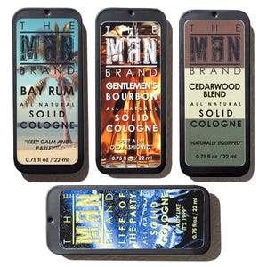 May include: An assortment of The Man Brand solid colognes in rectangular metal tins. The scents include Bay Rum, Gentlemen's Bourbon, Cedarwood Blend, and Life of the Party. Each tin is labeled with the scent name and the phrase "All Natural Solid Cologne."