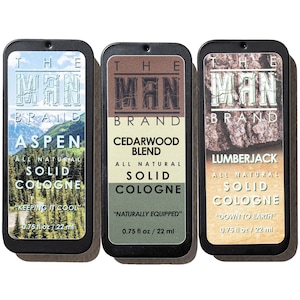 May include: Three rectangular tins of solid cologne from The Man Brand. The tins are black with labels featuring the brand name and scent names: Aspen, Cedarwood Blend, and Lumberjack. Each tin contains 0.75 fl oz / 22 ml.