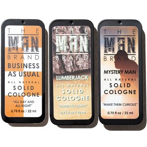 May include: Three rectangular solid cologne tins from The Man Brand. The tins are black with rounded corners and feature different designs and text. The first tin is orange and white, the second is brown and green, and the third is a sunset color.