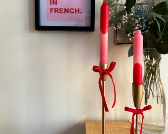 May include: Two gold candle holders with red ribbon bows and pink and red candles. The candles are on a wooden table. The text "I LOVE YOU. IN FRENCH." is on a pink background in a black frame on the wall.