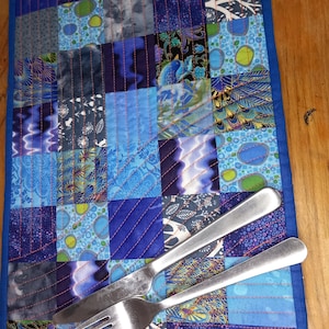 A vibrant blue patched patchwork fabric table mat with curved  and straight orange quilting, special occasion tableware, table linen.