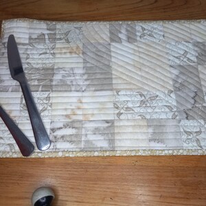 May include: A white quilted placemat with a geometric pattern and a subtle beige and brown colour scheme. Two silver utensils are placed on the placemat.