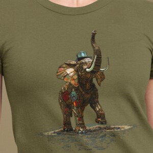 Peut inclure: T-shirt vert olive avec une illustration détaillée d'un éléphant portant un chapeau. L'éléphant est orné de motifs mécaniques et colorés complexes, avec sa trompe levée. Le motif est posé sur un fond subtil ressemblant à de l'eau.