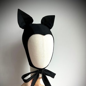 May include: Black cat-shaped hat with ears, made of fabric. The hat has a tie that can be tied under the chin. The hat has a small label with the text "MESS" on it.
